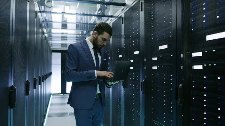 IT engineer dressed in blue suit working with laptop on managing hybrid cloud from server room.

