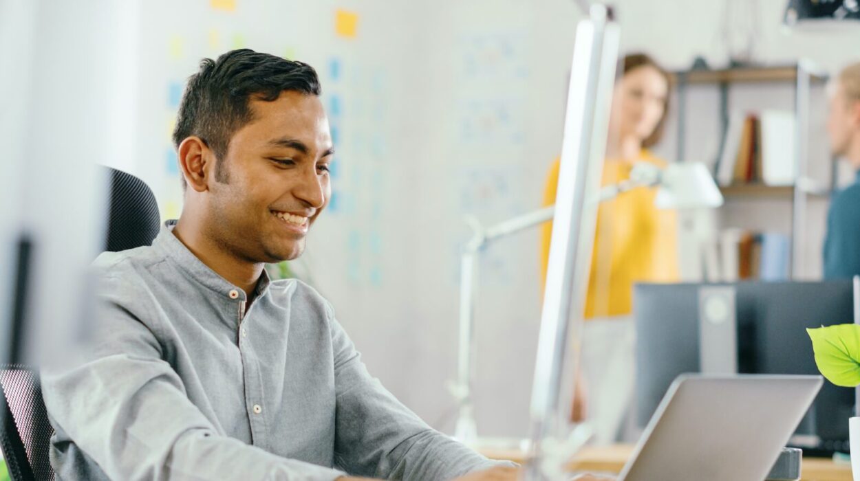 Handsome Smiling Indian Office Worker Sitting at His Desk works on a Laptop. In the Background Modern Office with Diverse Team of Young Professionals Working.