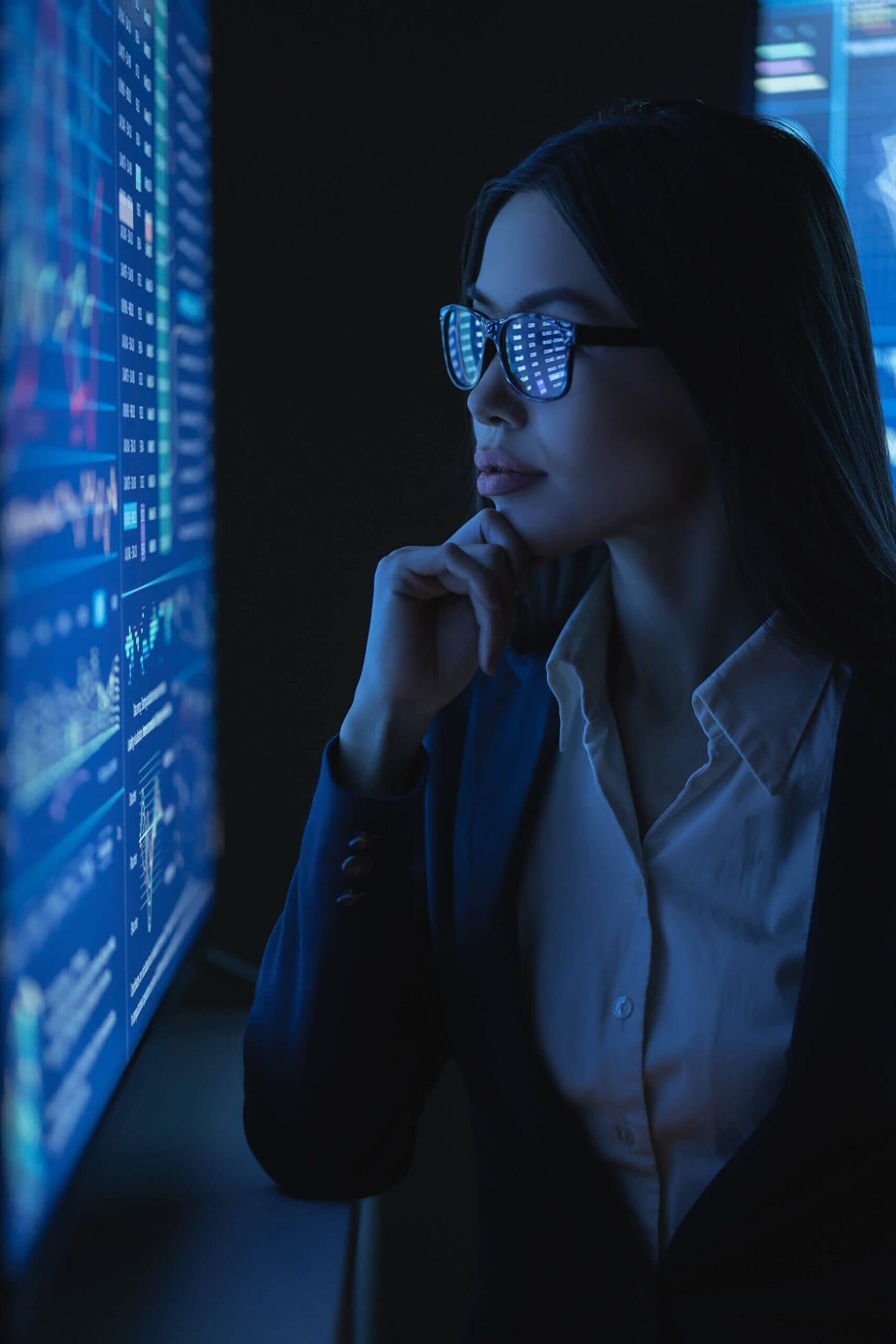Young female IT professional wearing glasses looking at Microsoft reports displayed on monitor.